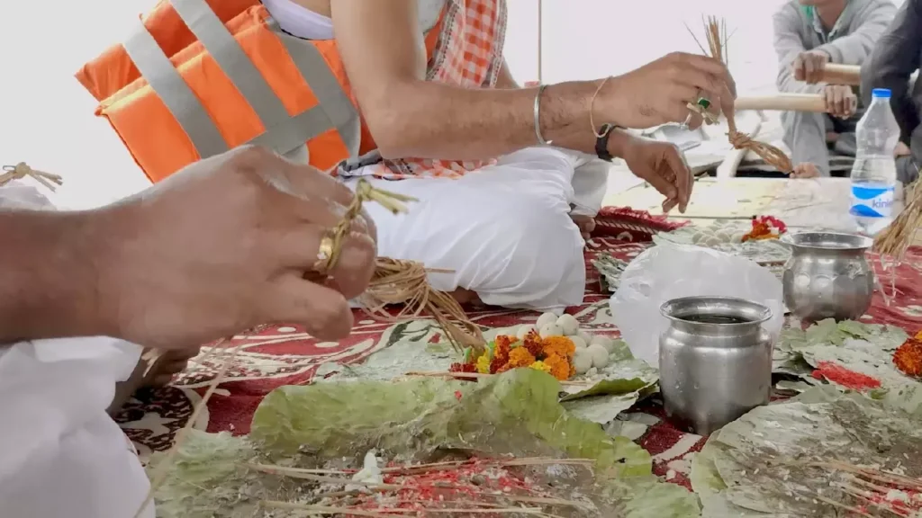 Men participate in a traditional ritual aboard a boat. Dressed in customary attire, with one wearing an orange life vest, they handle offerings like flowers and food placed on leaves. Another man rows the boat in the background during what appears to be a riverside ceremony.