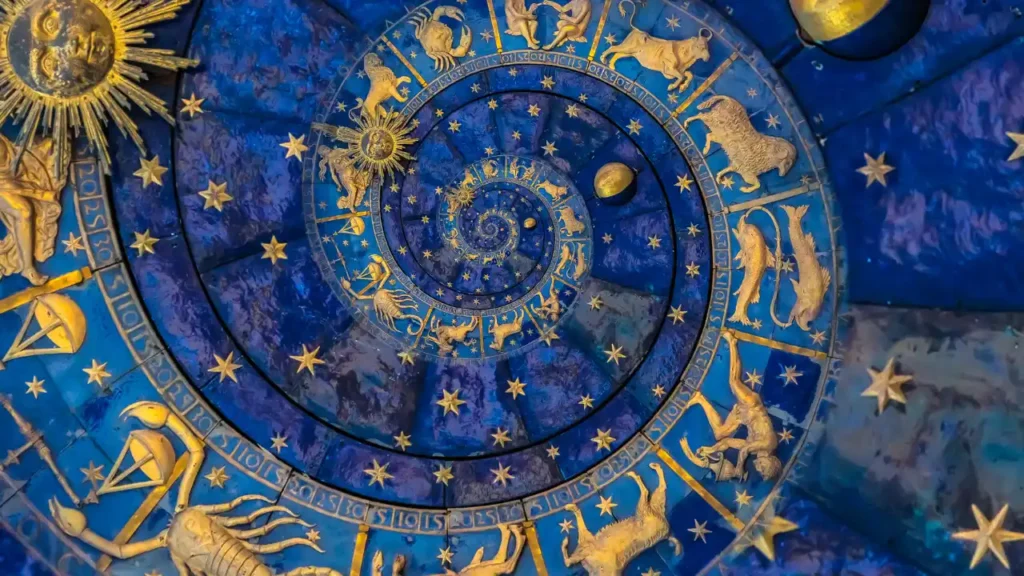 The image shows a detailed illustration of a zodiac wheel or astrological chart designed as a spiral. It's rendered in rich, deep blues and accented with elaborate gold details. Golden representations of the twelve zodiac signs (like Aries the ram, Taurus the bull, Gemini the twins, etc.) are clearly visible, arranged along the spiral path. Numerous golden stars are scattered across the blue background. A prominent golden sun symbol is visible towards the outer edge, and another smaller golden sphere (perhaps the moon or a planet) is near the center. The overall impression is one of ancient astronomy, astrology, or a celestial clock face.