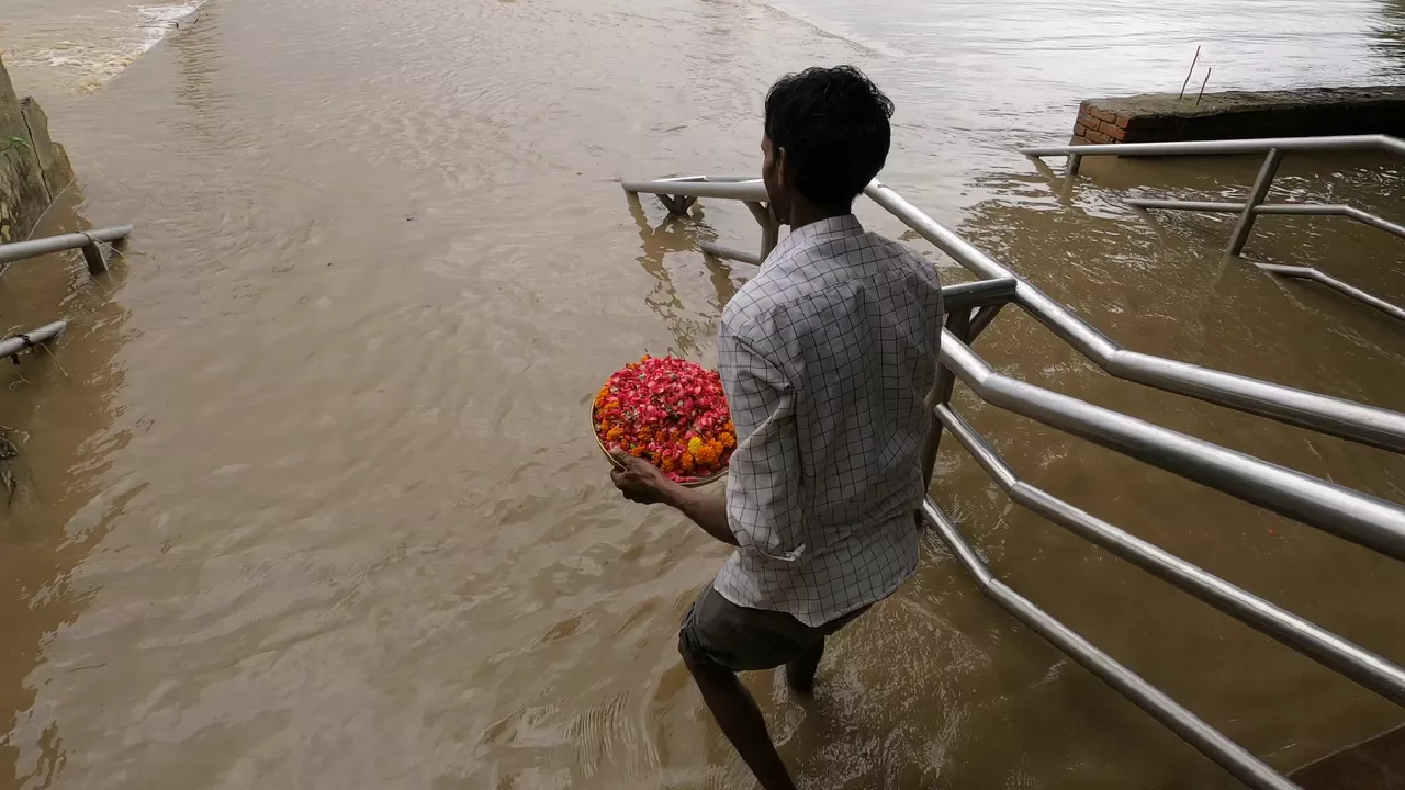 Man carries a plate of flower petals into a flooded river, standing on submerged steps for offering.
